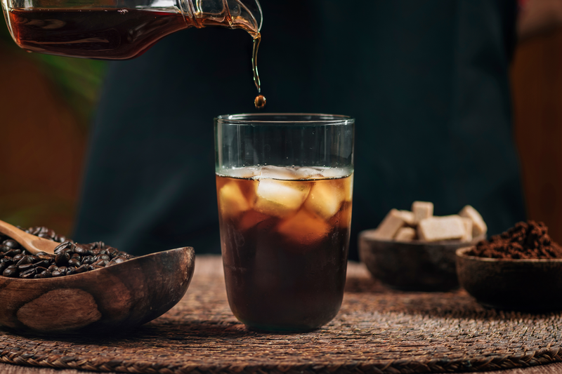 Glass of iced coffee being poured with coffee beans and sugar cubes on a wooden surface.